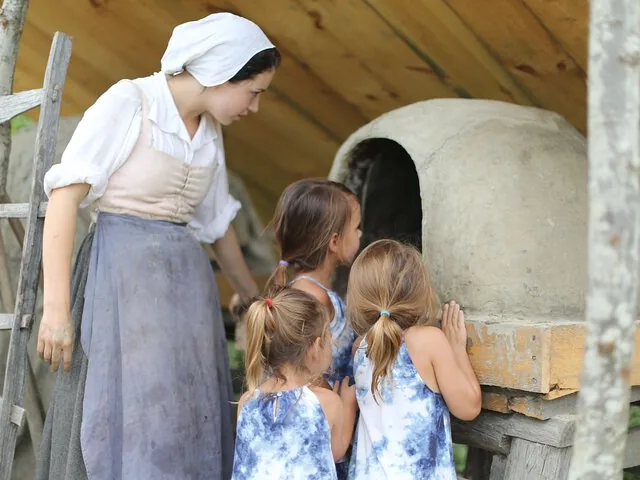 A woman in historical dress shows an outdoor oven to children.