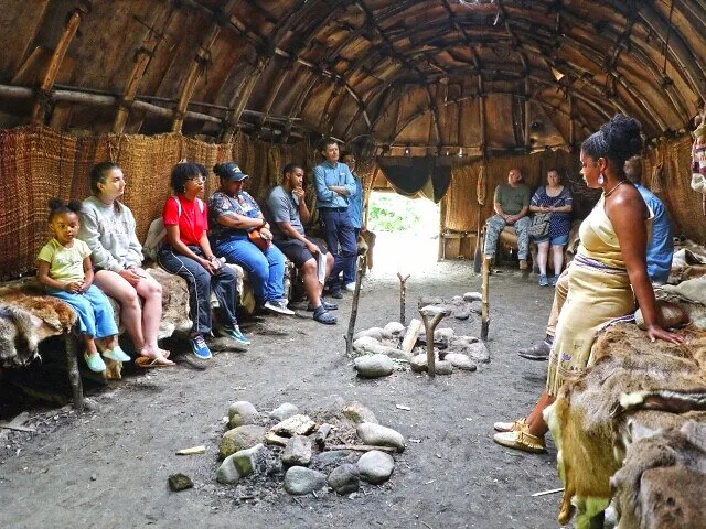 A group of tourists sit inside a Native American dwelling.